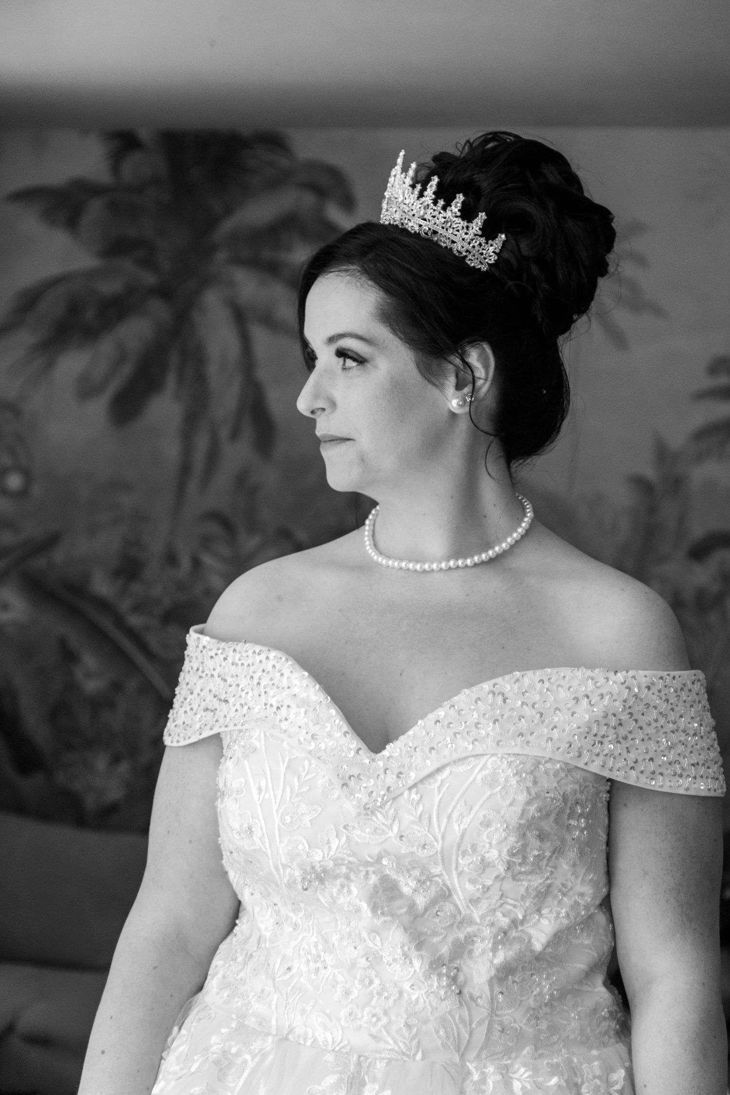 Black and white portrait of a bride wearing a tiara, pearl necklace, and off-shoulder lace wedding gown.