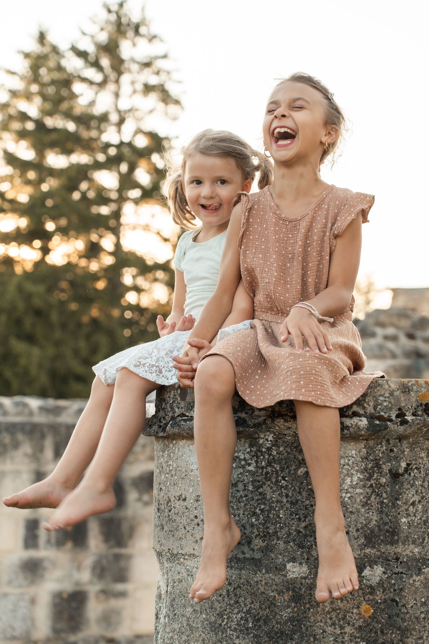 Two barefoot sisters in summer dresses laugh joyfully while sitting on a stone wall outdoors.
