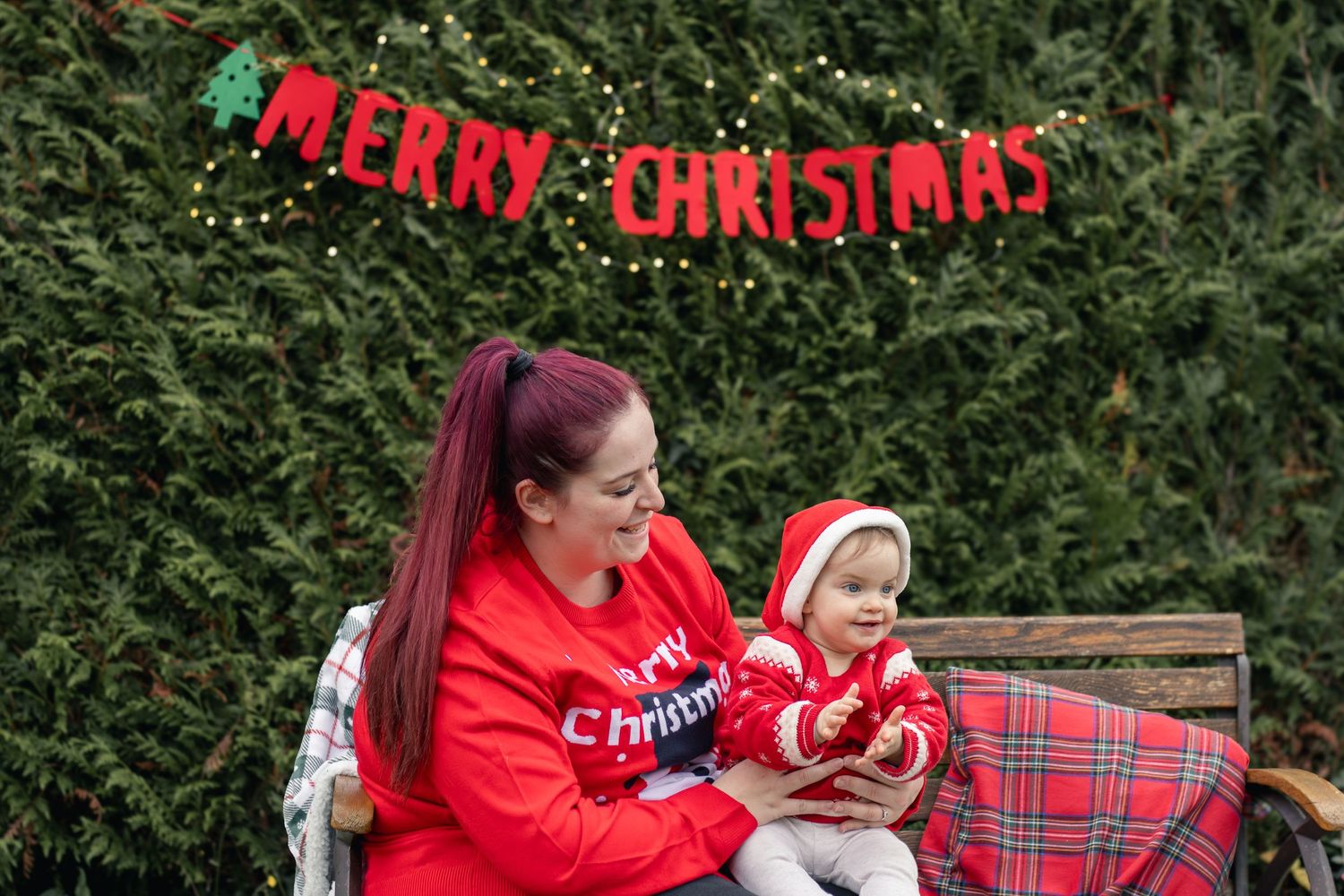 A mother in a red Christmas sweater holds her baby near a Merry Christmas banner outdoors.
