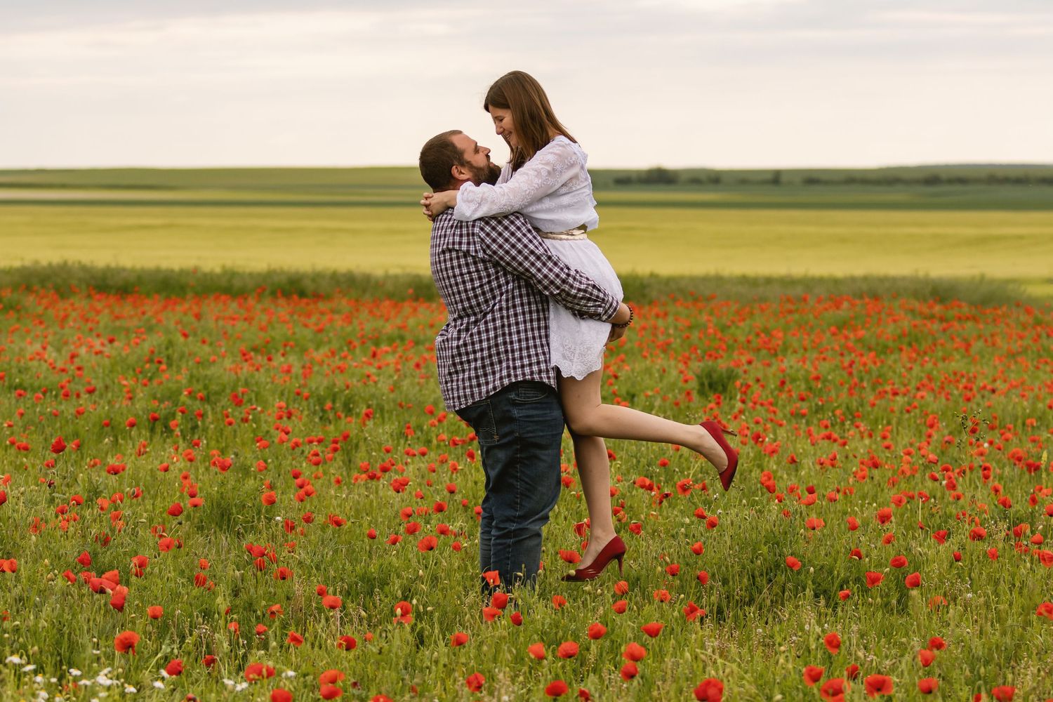 Couple embracing in a vibrant red poppy field, man lifting woman as they share a romantic moment outdoors.