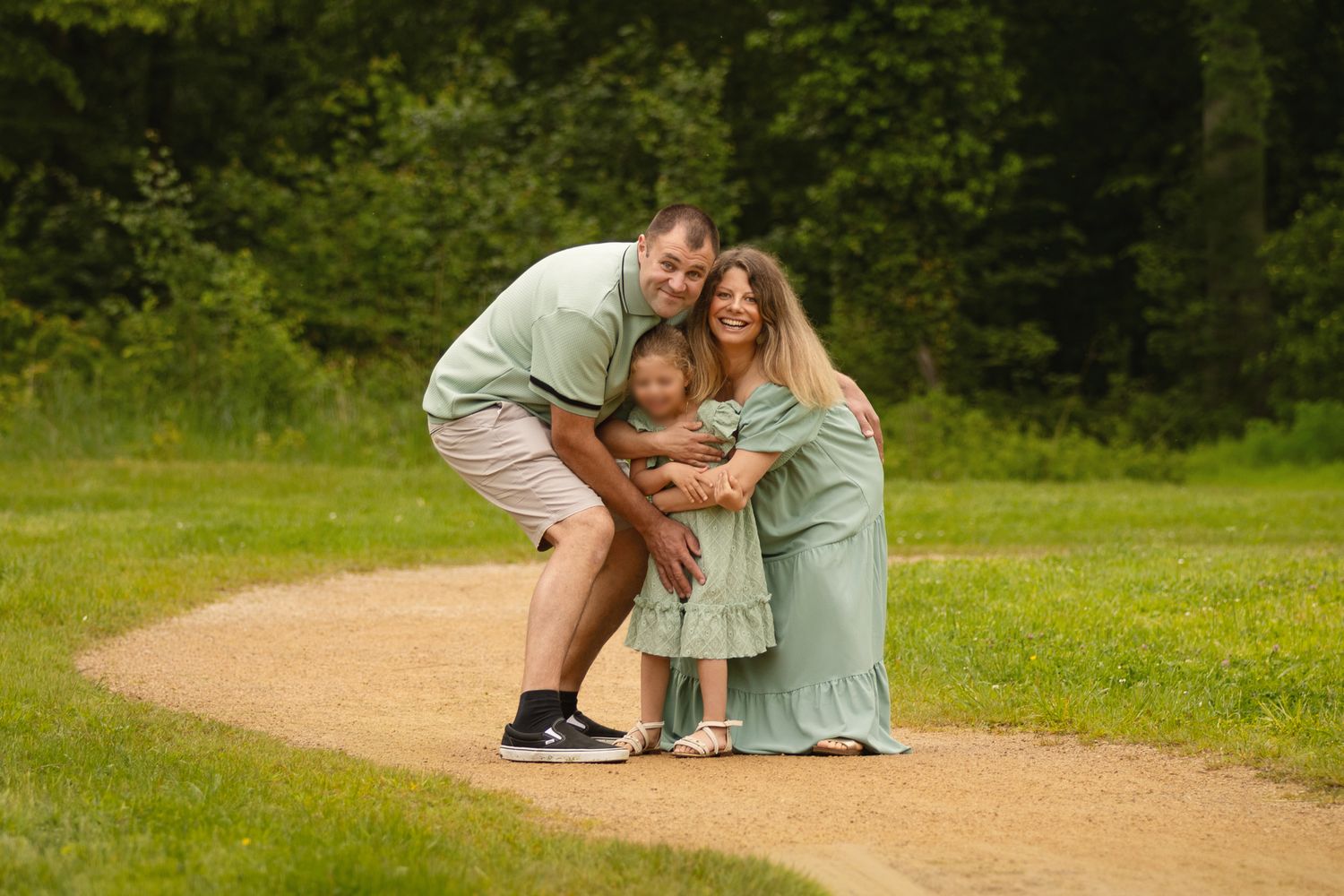 Family of three in matching green outfits posing on a garden path surrounded by lush trees.