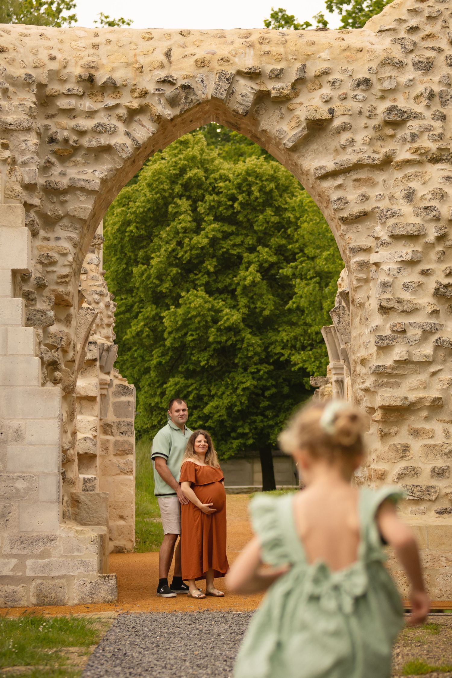 Pregnant couple poses under a stone arch ruin in a park, with a child in a green dress in the foreground.