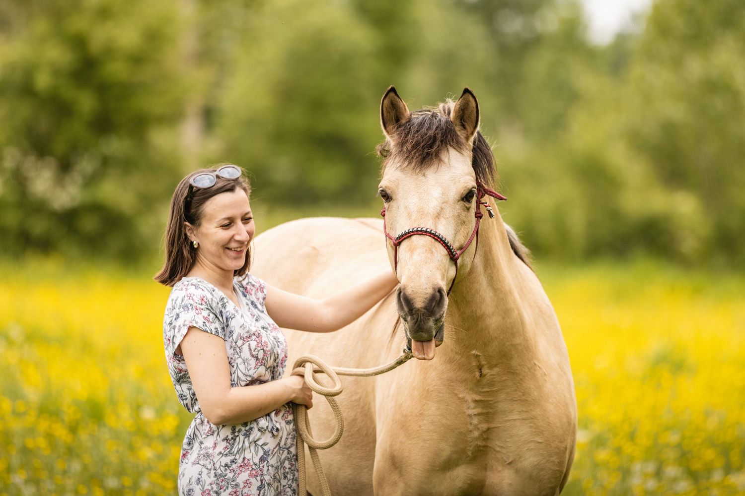 A smiling woman in a floral dress holds a rope lead while standing beside a palomino horse in a yellow wildflower field.