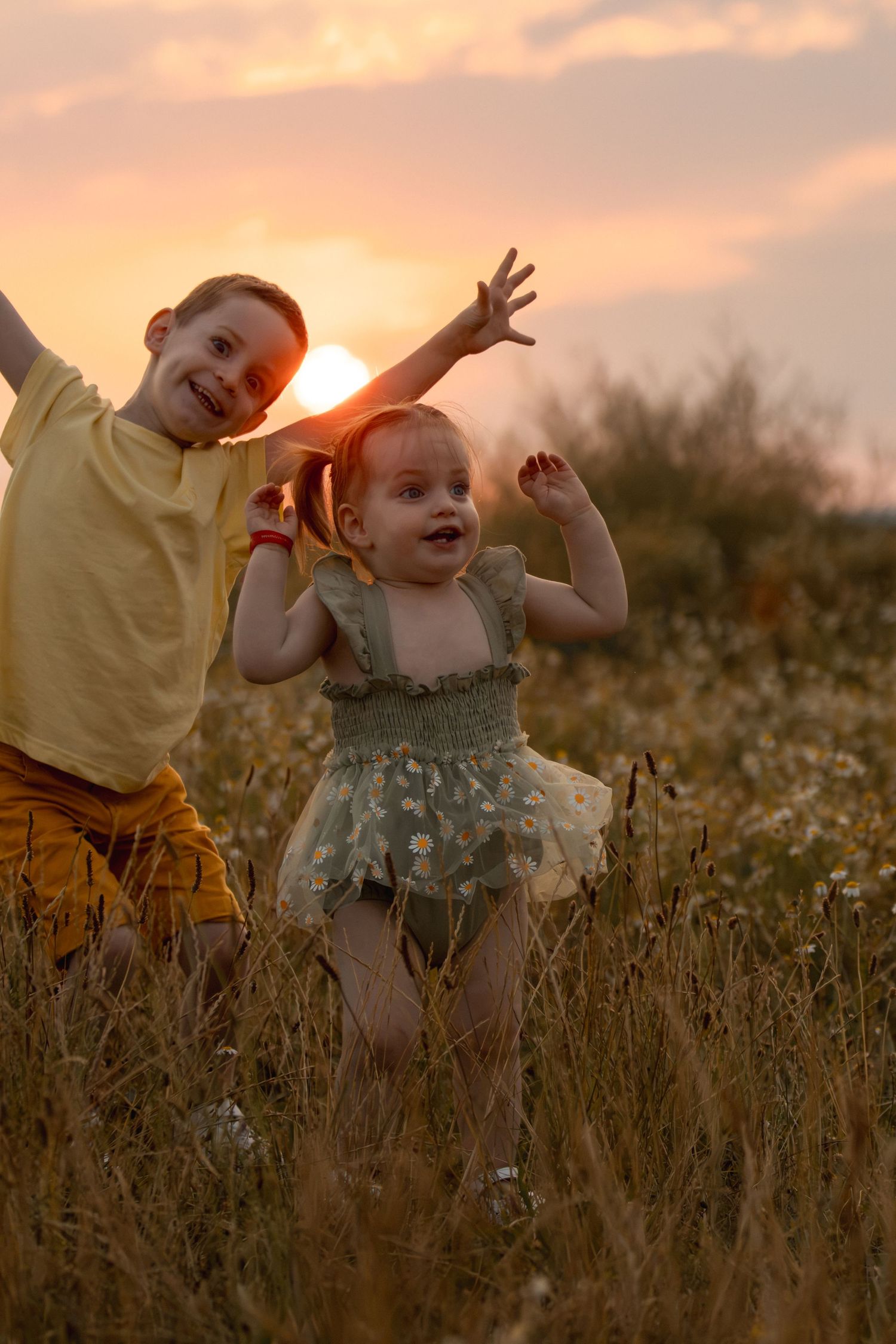 Two joyful children playing in a golden sunset meadow, boy in yellow shirt and toddler girl in floral dress with arms raised.