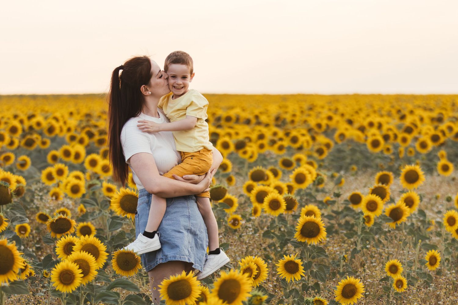Mother kissing her toddler son while standing in a vibrant yellow sunflower field at sunset.