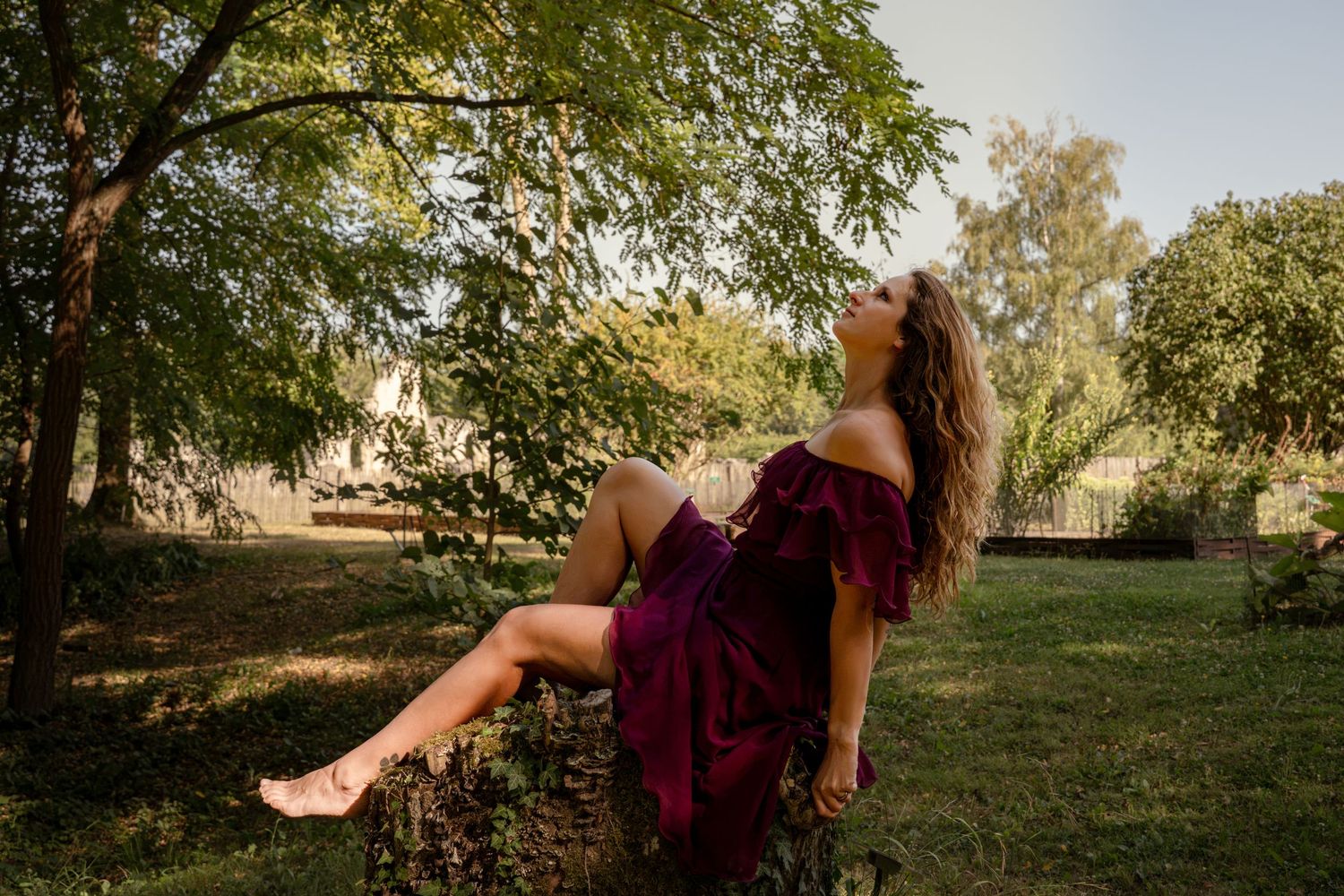 Woman in burgundy dress sitting on a tree stump, head tilted back, surrounded by lush green trees in a sunny garden.