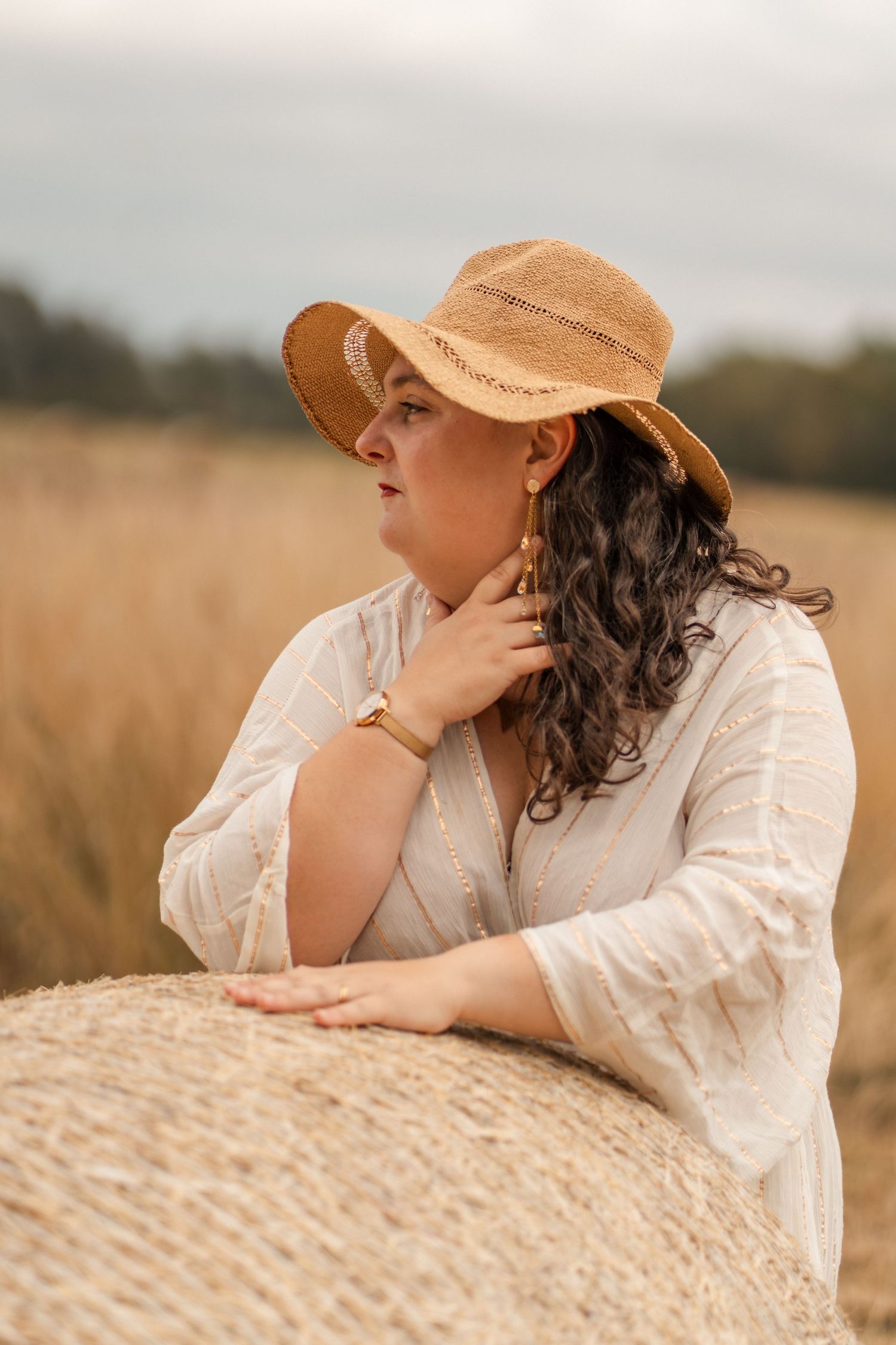 Woman in straw hat and white dress leaning on hay bale in golden field, wearing gold jewelry.