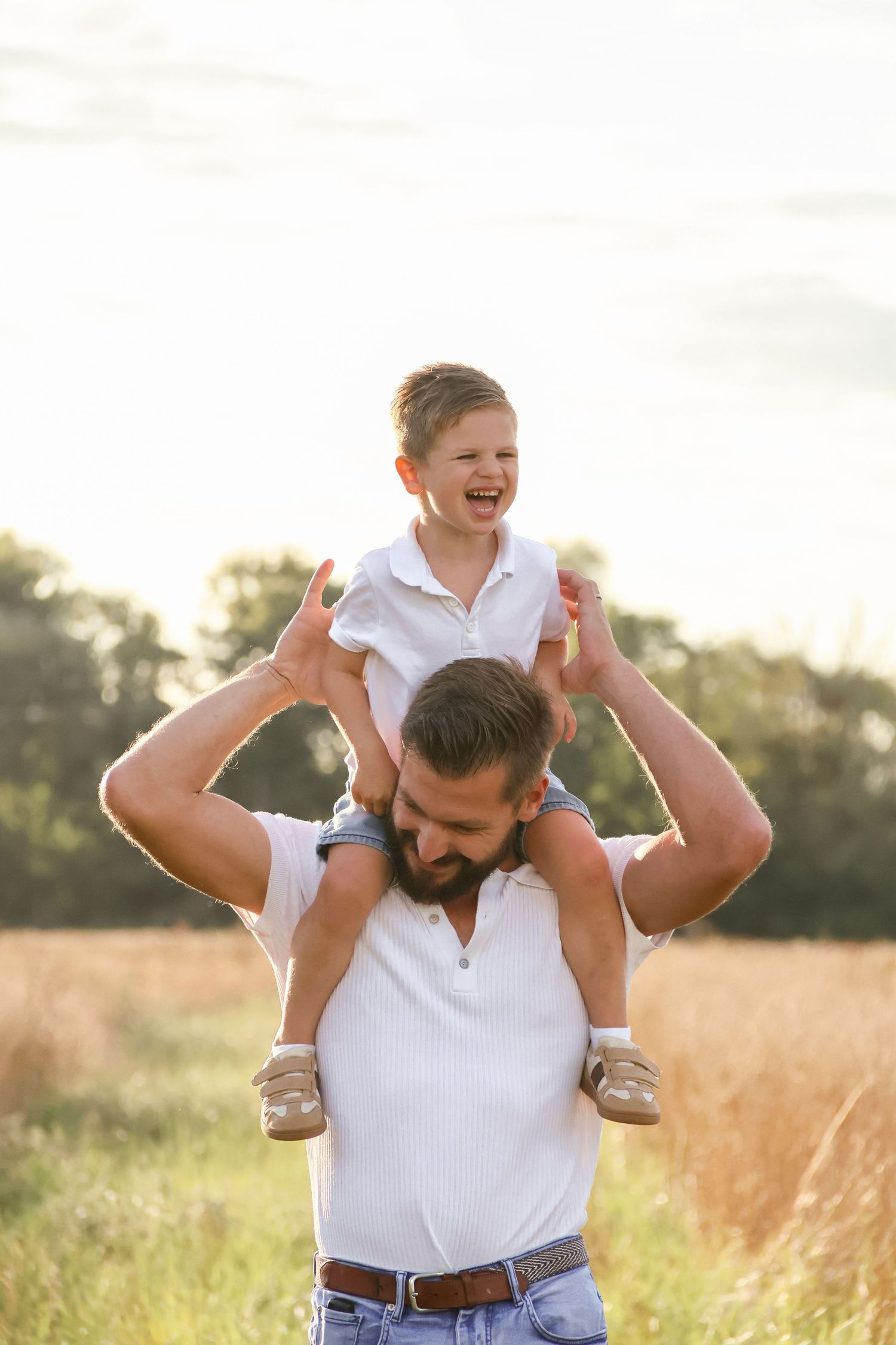 Laughing child sitting on father's shoulders in a sunlit field during golden hour.