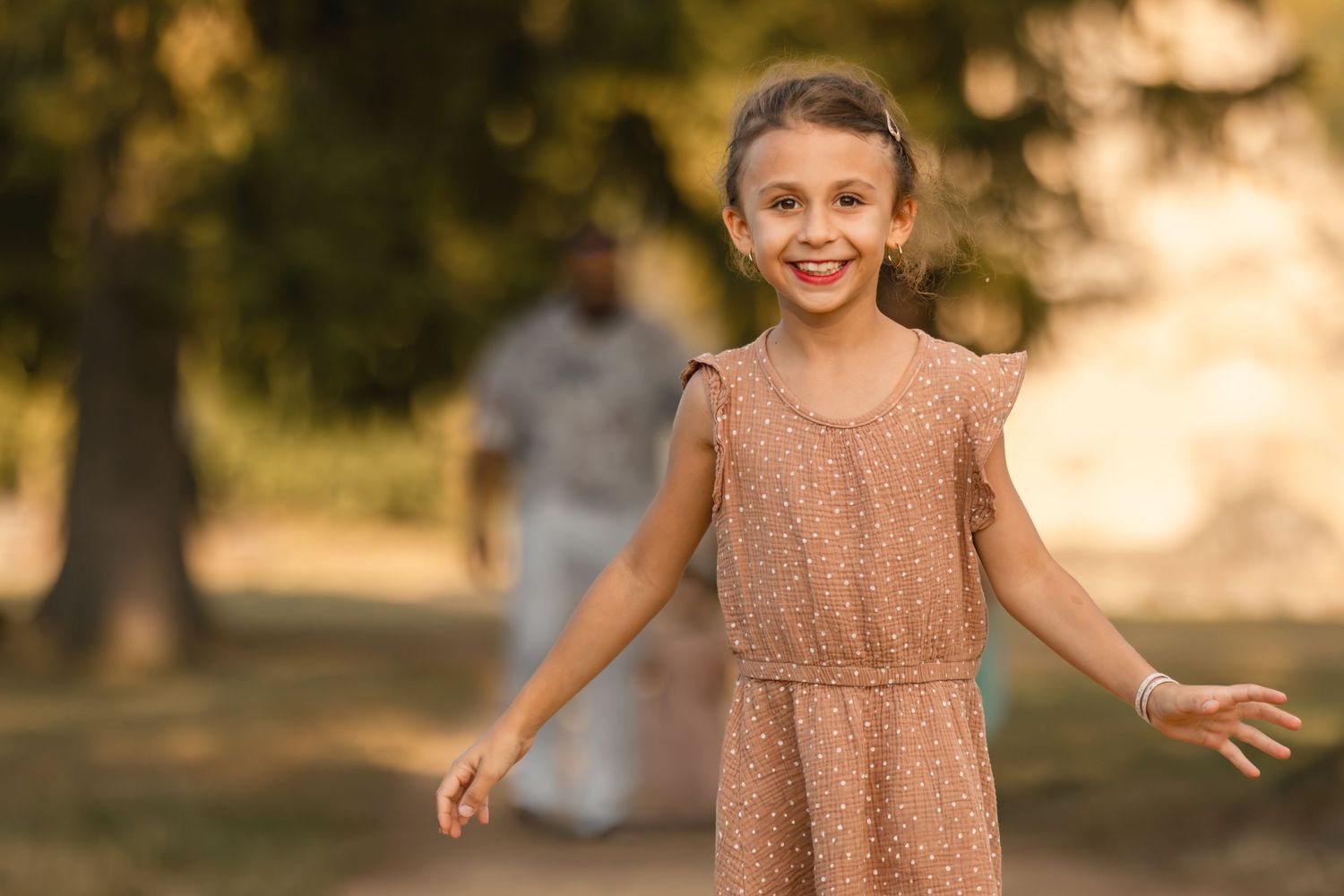 A happy young girl in a pink polka dot dress laughs with arms outstretched in a sunny park.