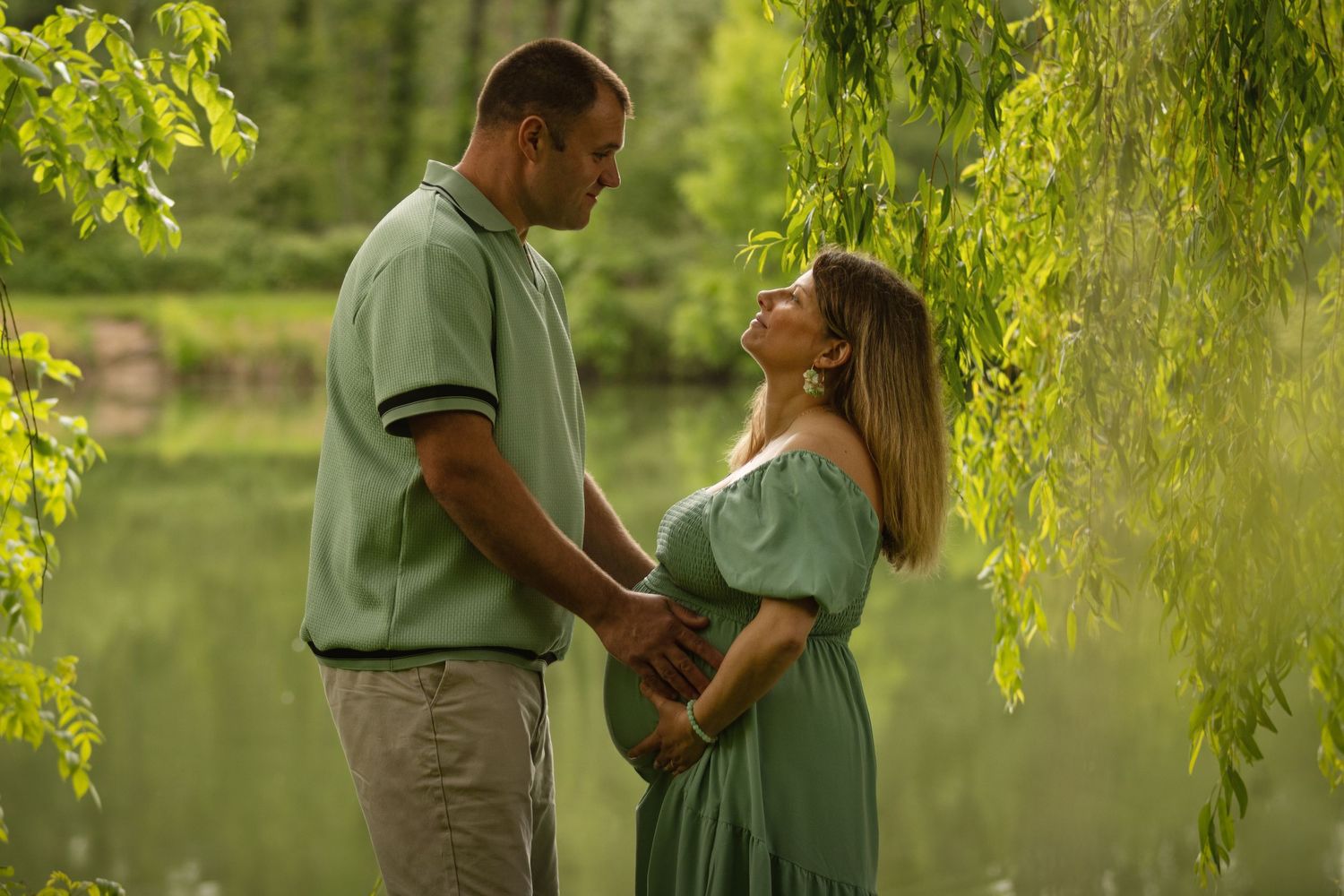 Couple holding hands by a pond under willow trees, woman in green maternity dress during outdoor pregnancy photoshoot.