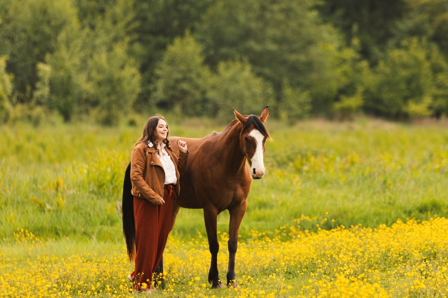 Woman in brown jacket stands beside a chestnut horse in a sunny field of yellow wildflowers with green trees behind.
