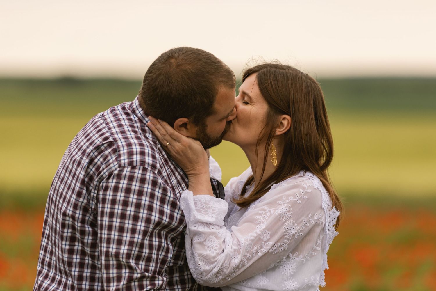 A couple shares a romantic kiss in a blooming poppy field, the woman in white and man in plaid shirt.