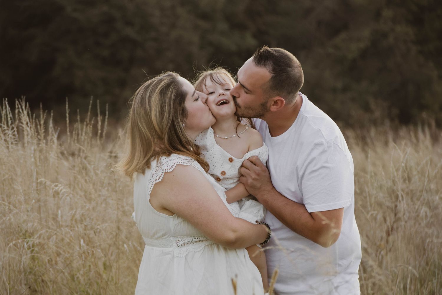 Family of three sharing a joyful moment in a golden field, parents kissing their young child on the cheeks.