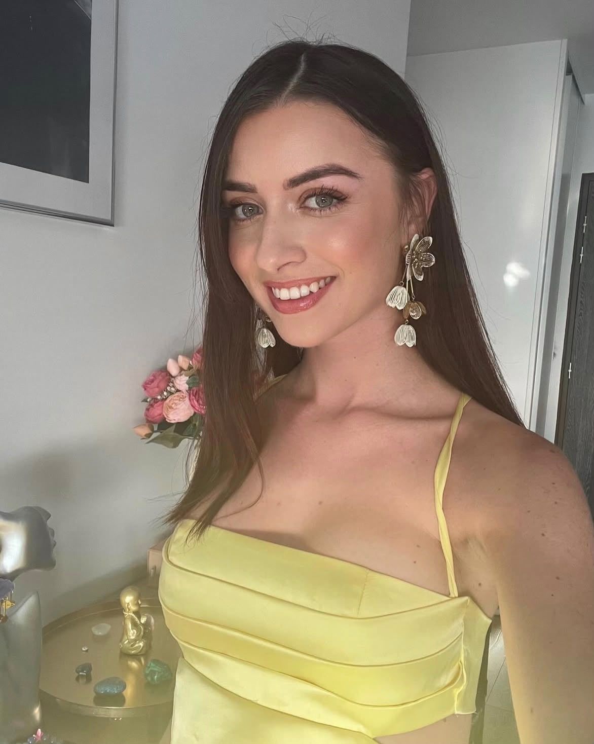 Young woman in yellow satin dress with gold earrings smiling for a selfie with pink flowers in background.