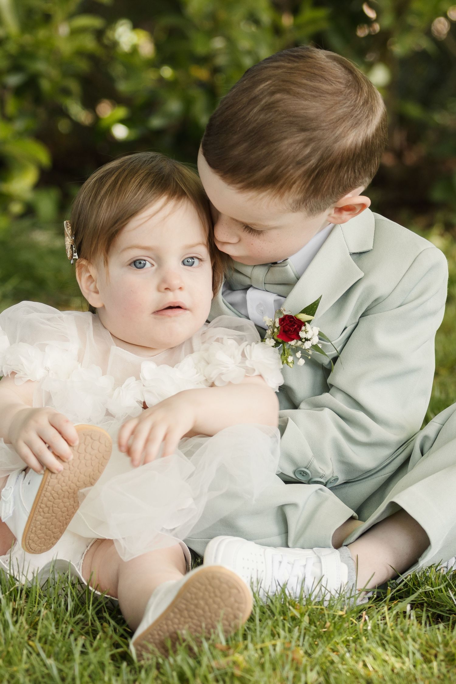 A young boy in a sage suit with boutonniere kisses a baby girl in a white dress while sitting on green grass.