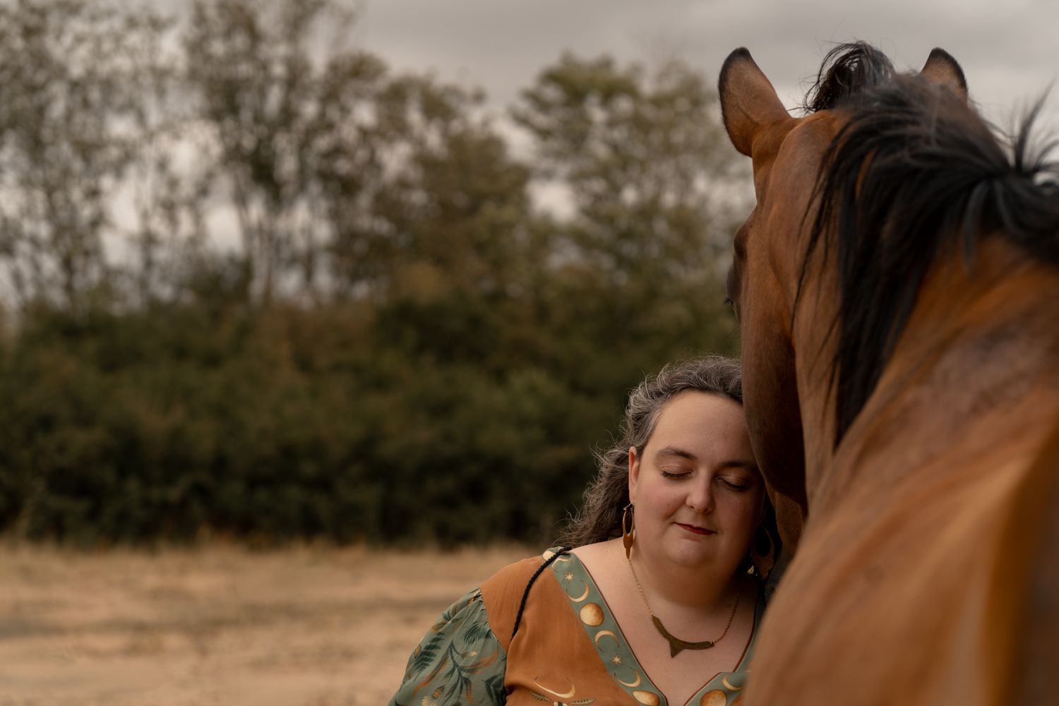 Woman in floral dress shares tender moment with chestnut horse in a rural field under overcast skies.