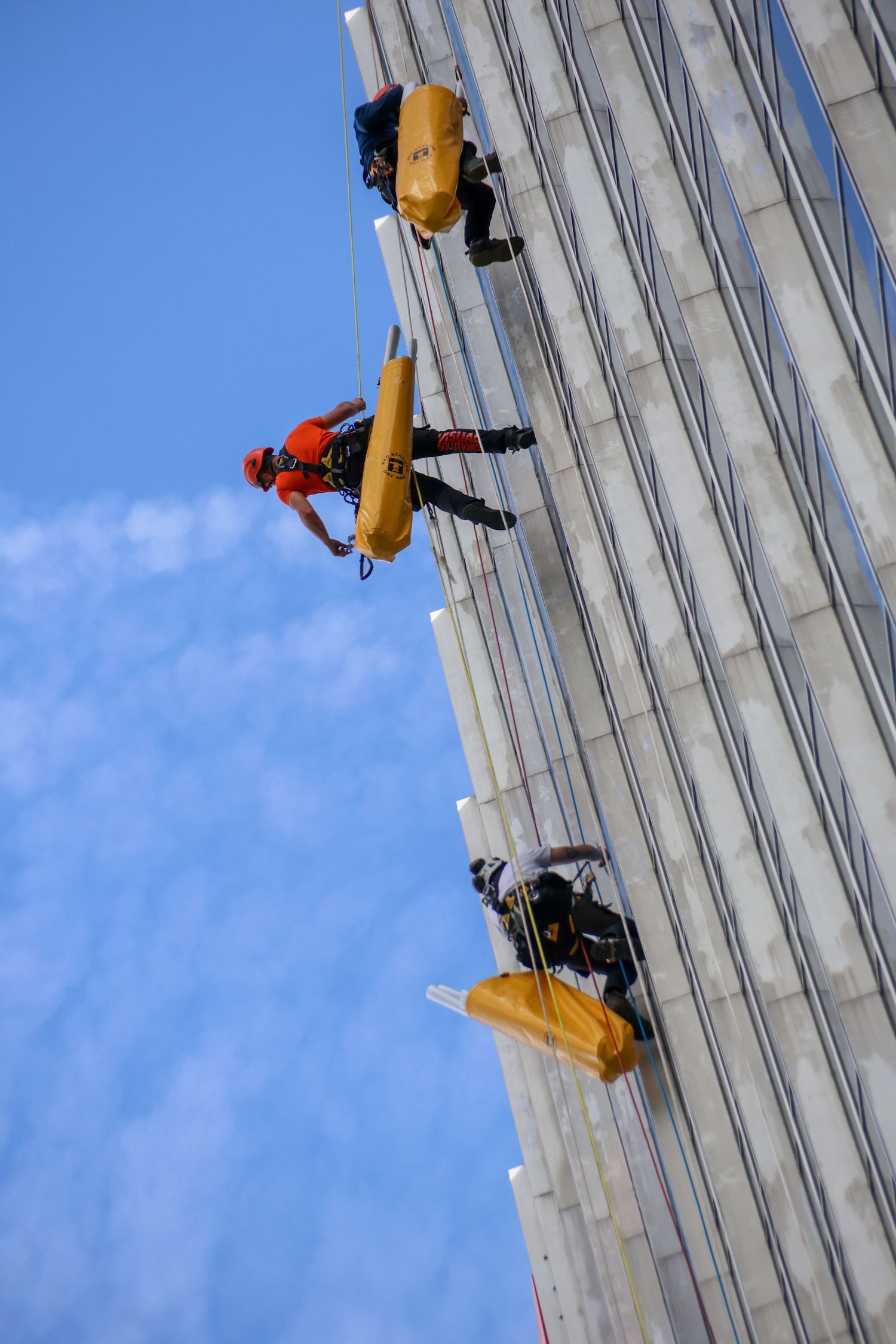 Three workers rappelling down a tall skyscraper facade, wearing safety harnesses and helmets against a blue sky.