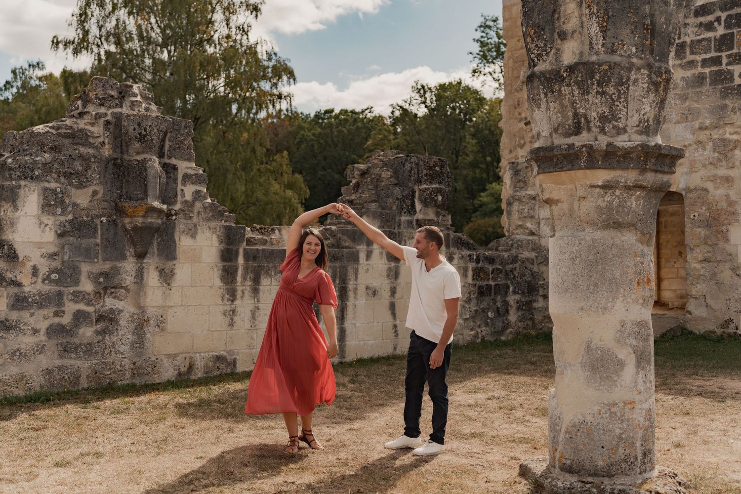 Couple dancing among ancient stone ruins, woman in red dress, man in white shirt, lush greenery backdrop.