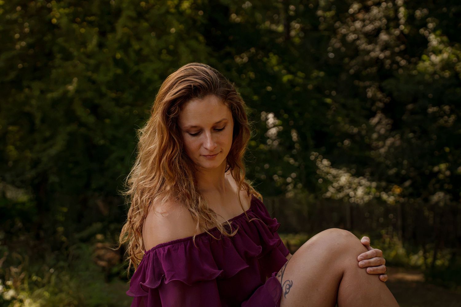 Woman in purple off-shoulder dress sitting outdoors with lush green trees in background.