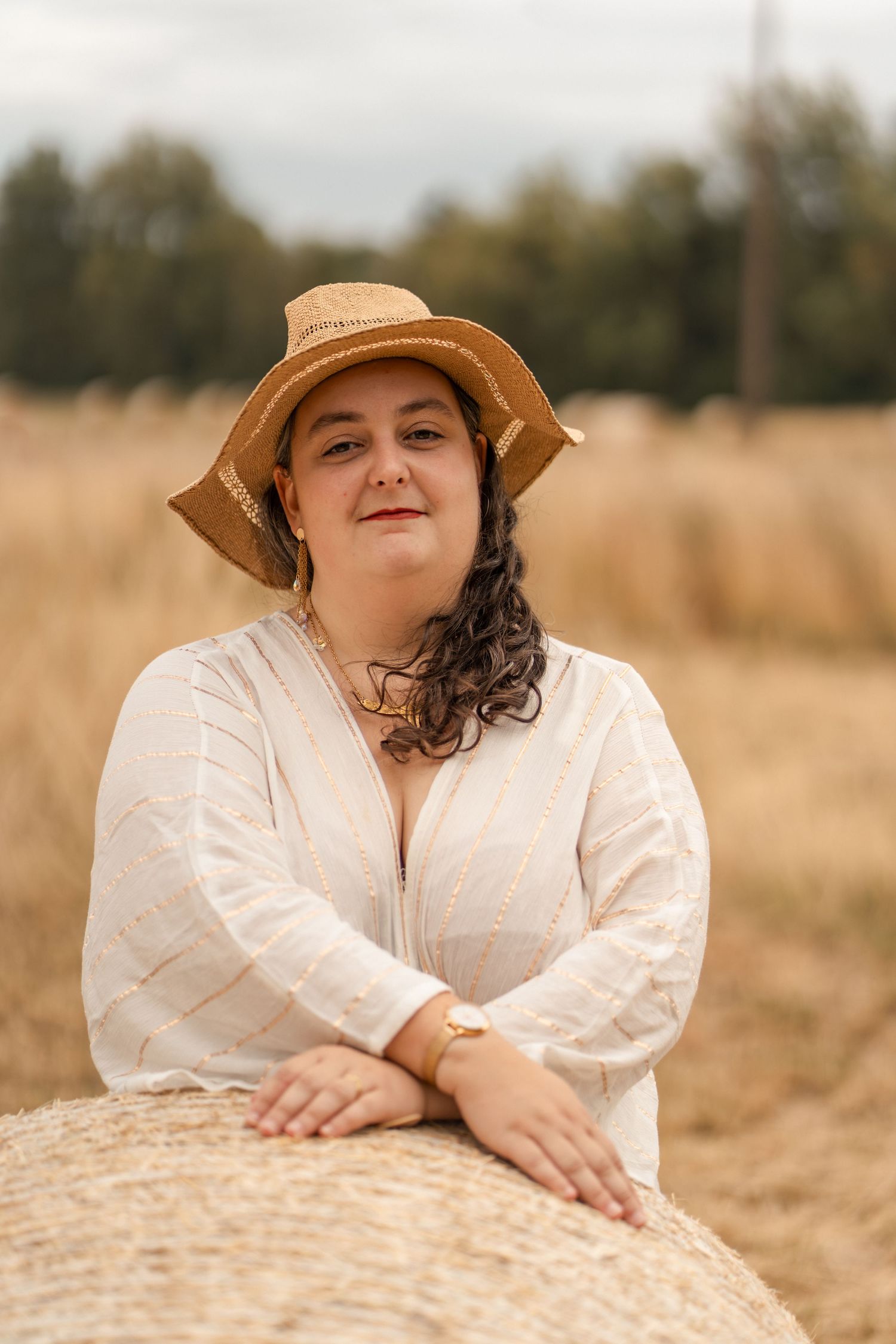 Woman in white linen dress and straw hat leaning on hay bale in open field.