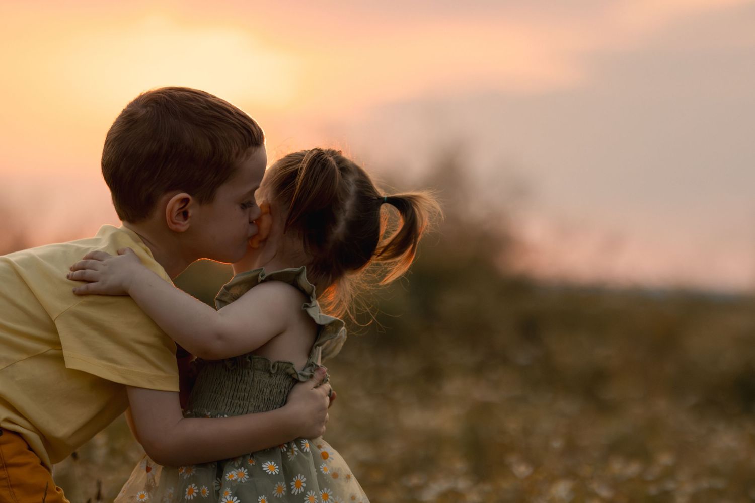 Two young children sharing a sweet kiss at sunset in a golden field, capturing an innocent and heartwarming moment.