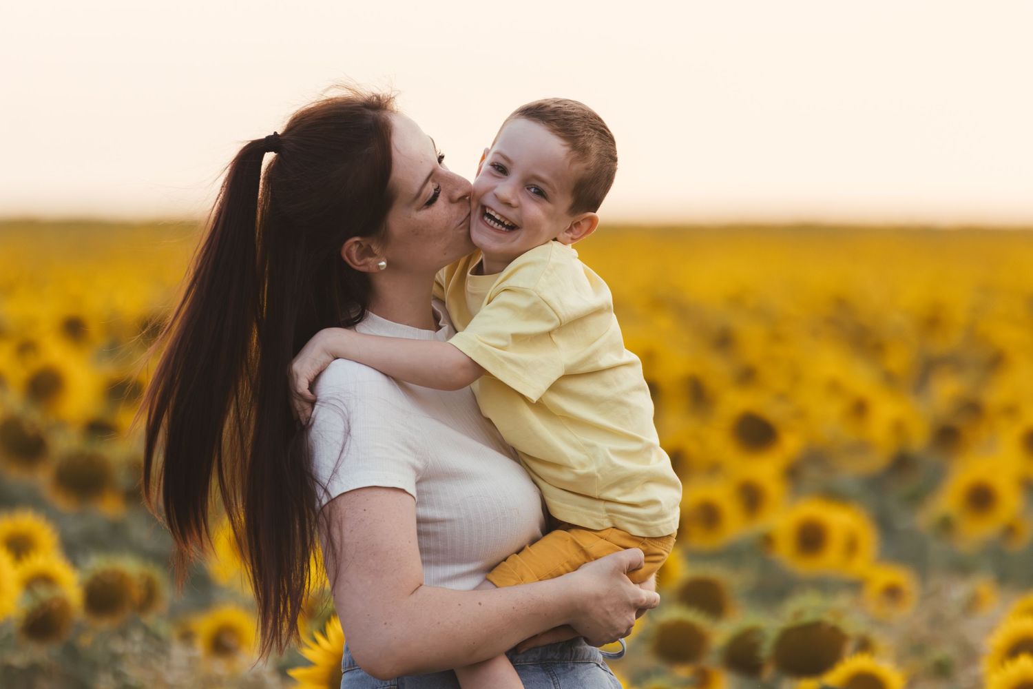 Mother kissing happy toddler boy in sunflower field at sunset.