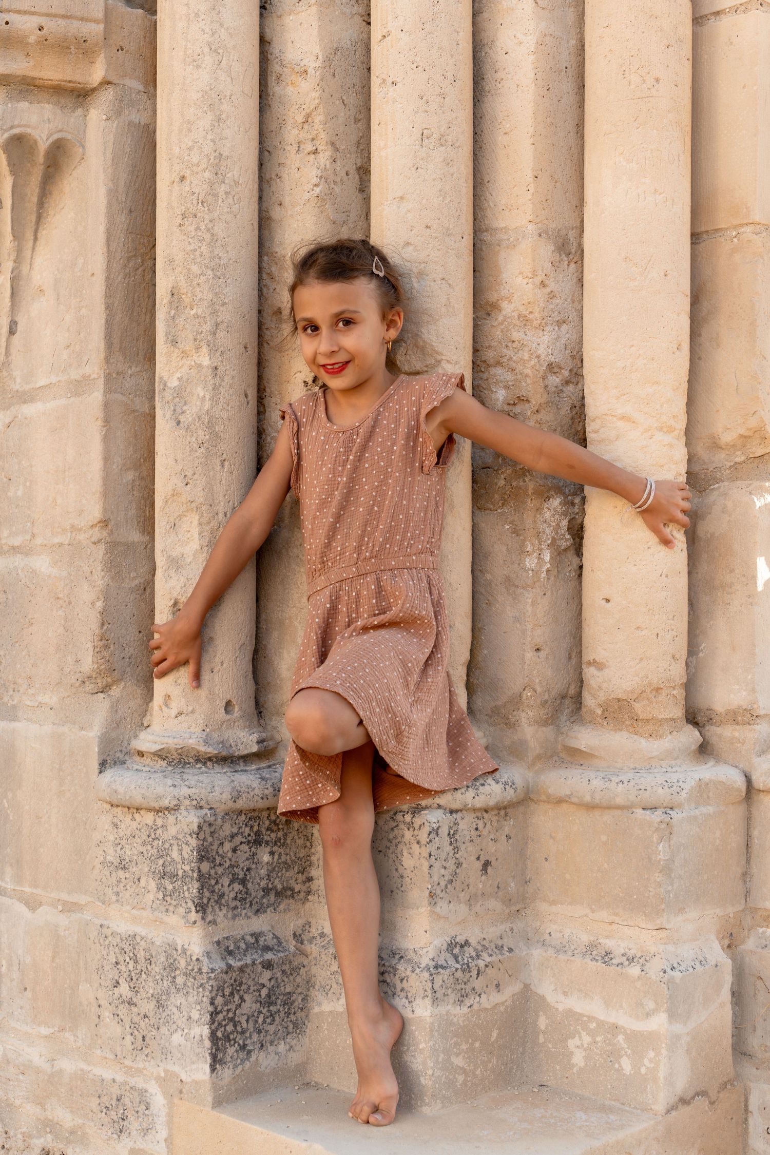 Barefoot girl in brown polka dot dress posing between stone columns of historic ruins.
