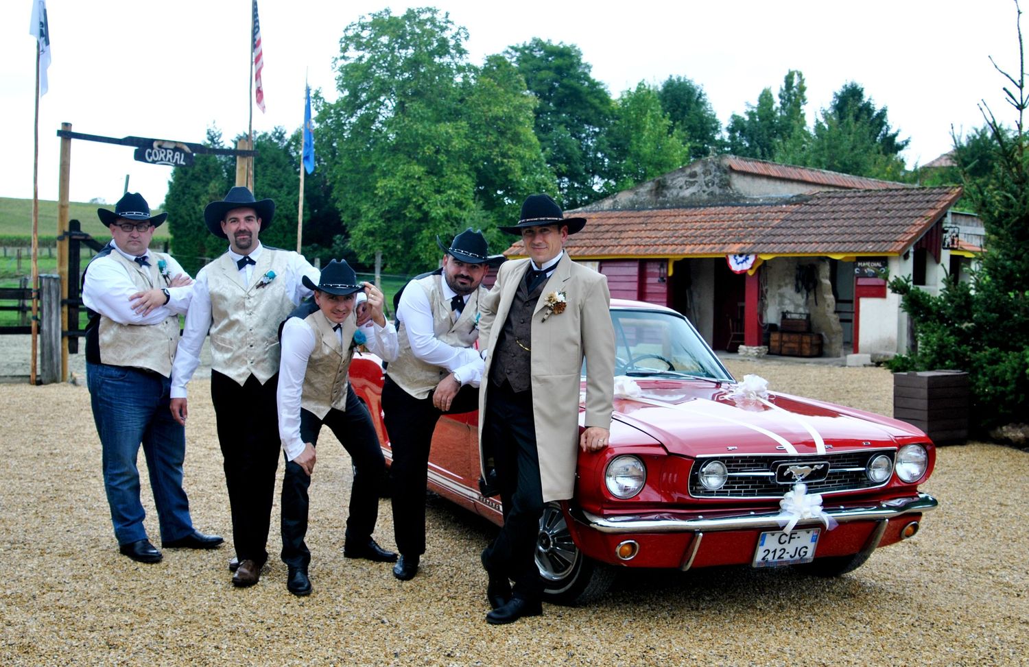 Four groomsmen in western vests and cowboy hats pose beside a classic red Ford Mustang at a western-themed wedding.