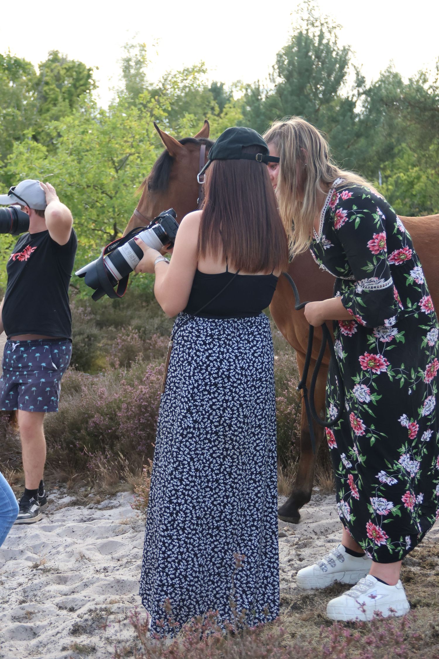 Two women photographing a brown horse outdoors near sandy terrain and trees.