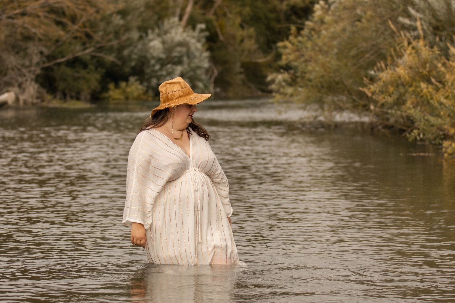 Pregnant woman in white lace dress and straw hat standing in a serene river surrounded by lush green trees.