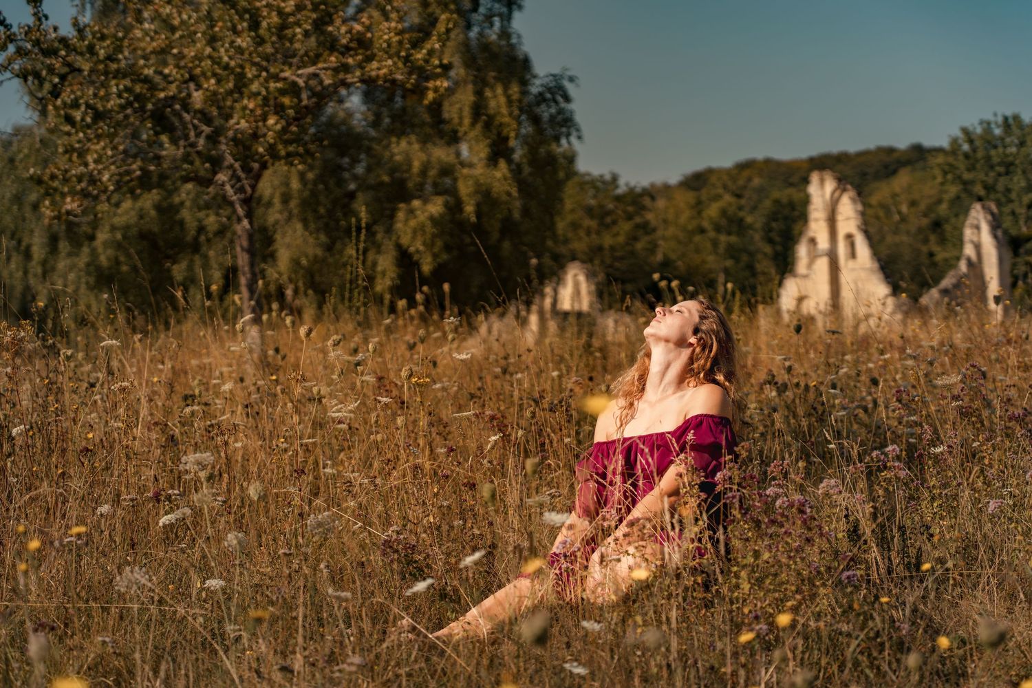 Woman in floral dress sitting in golden meadow, head tilted back, with ancient ruins and trees in background.