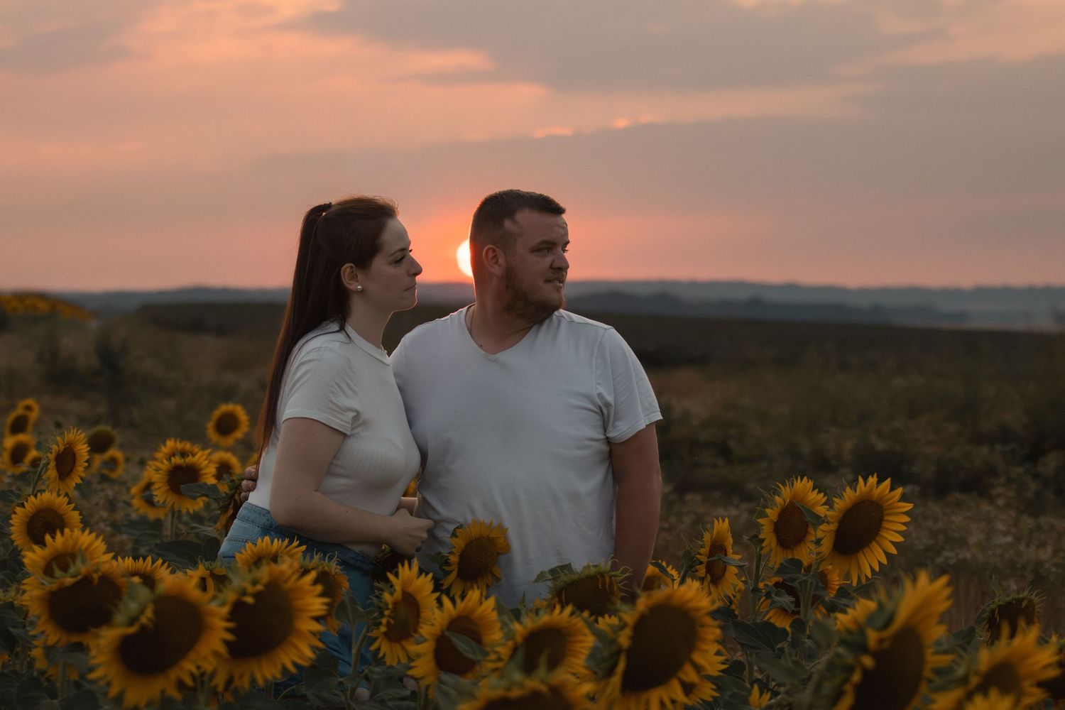 Couple standing together in a sunflower field at sunset, wearing white t-shirts, with a colorful orange sky in the background.