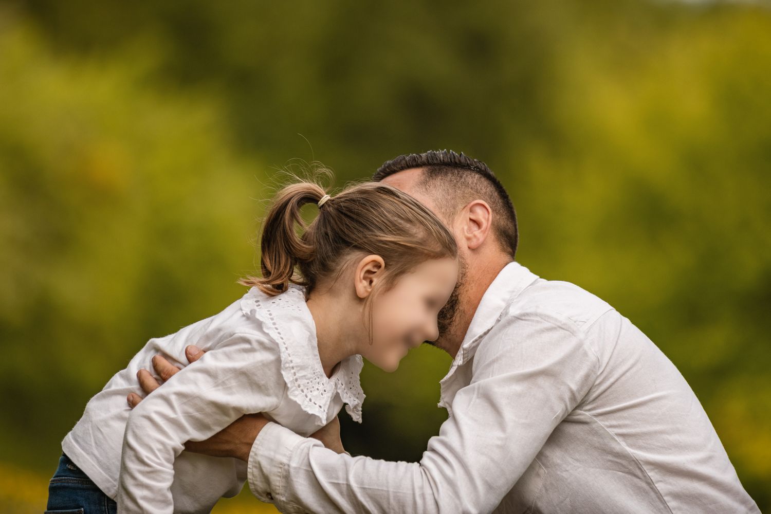 Father and daughter touching foreheads affectionately outdoors with green foliage background.