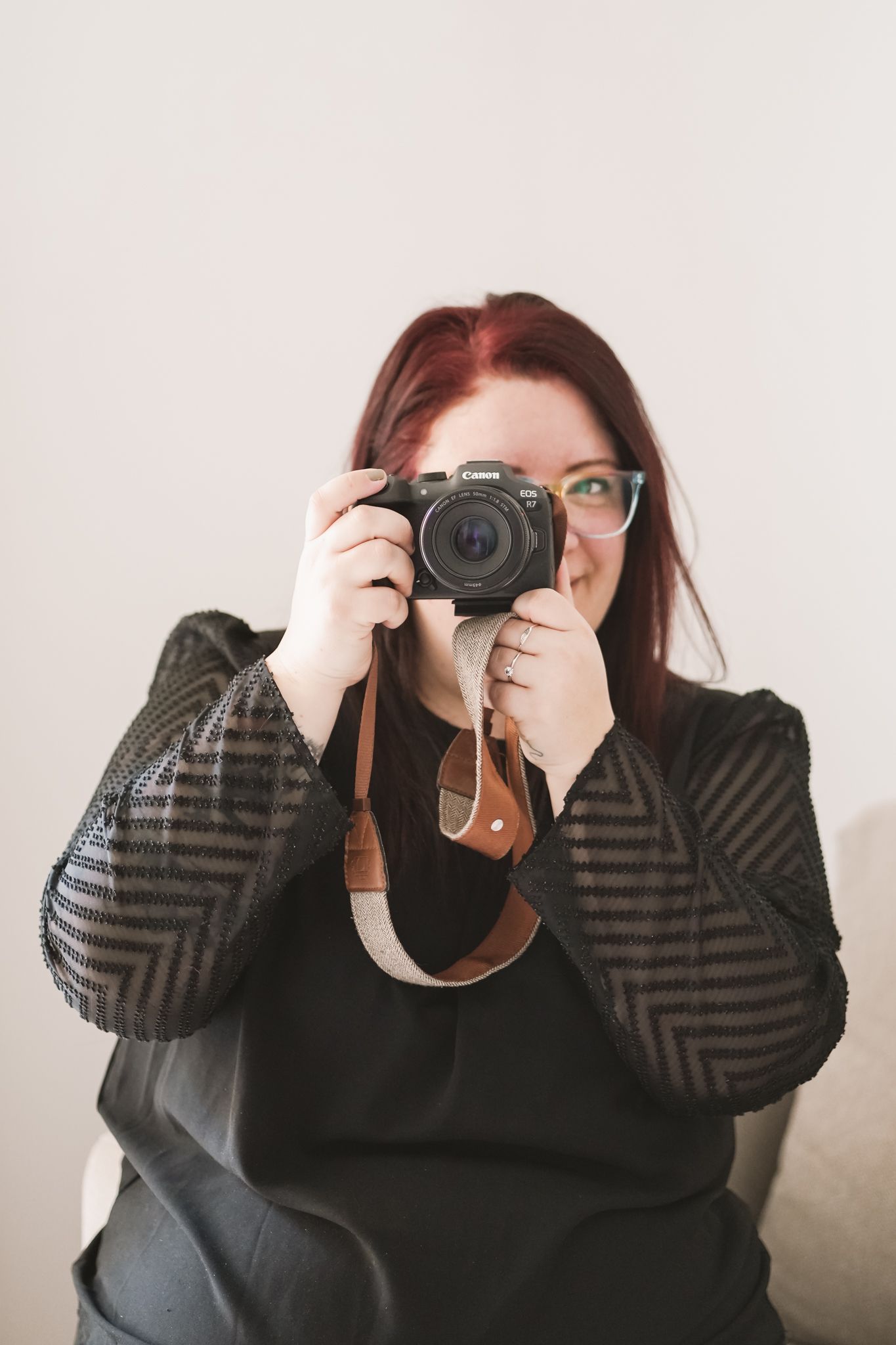 Woman with red hair holding a Canon camera up to her face, wearing a black outfit, seated against a light background.