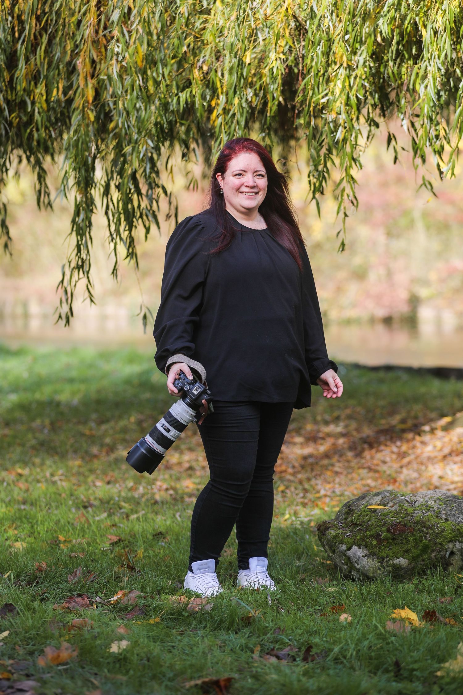 A smiling redhead woman in black outfit holds a professional camera with telephoto lens while standing under a willow tree.