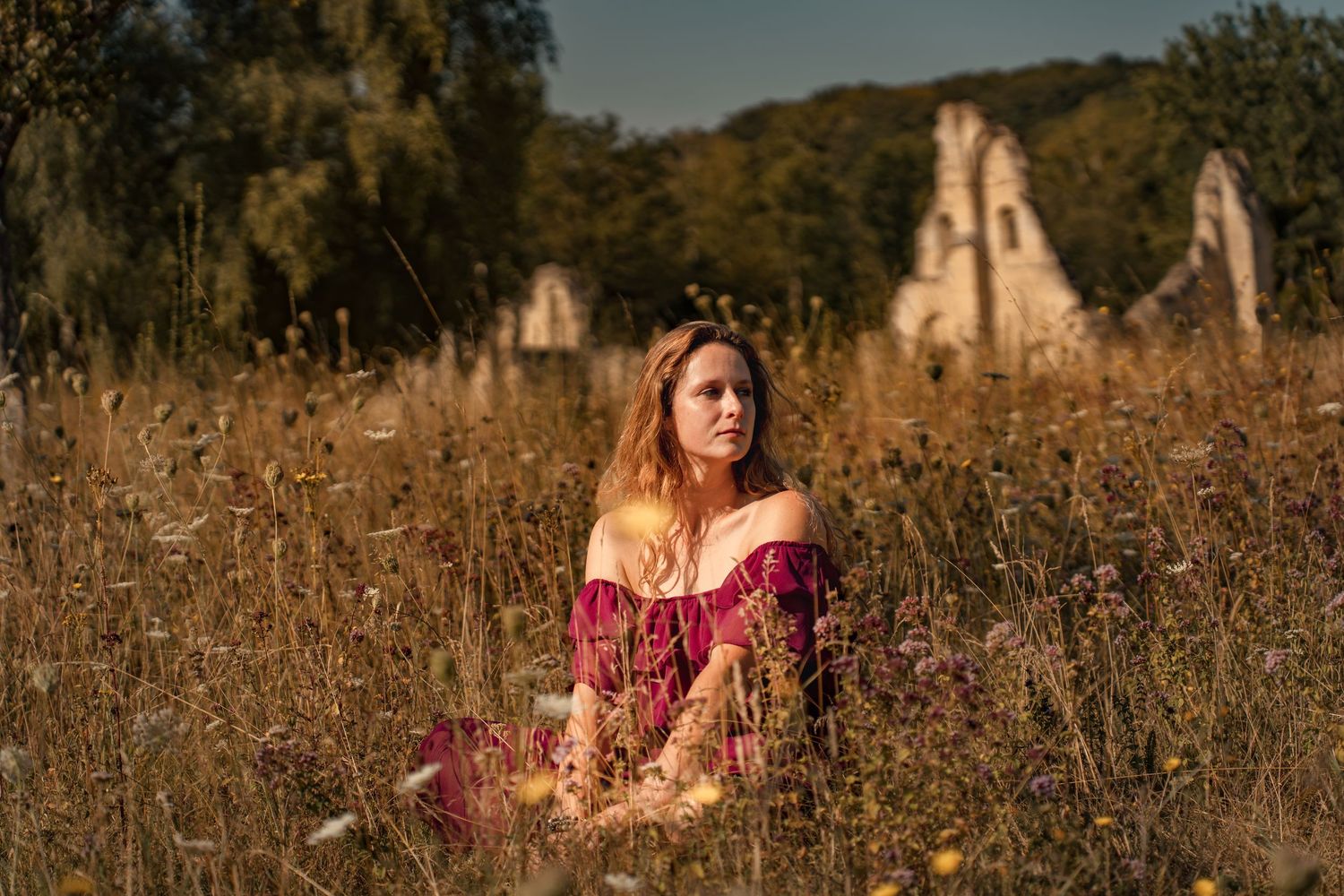 A woman in a red off-shoulder dress sits in a wildflower field with ancient stone ruins in the background.