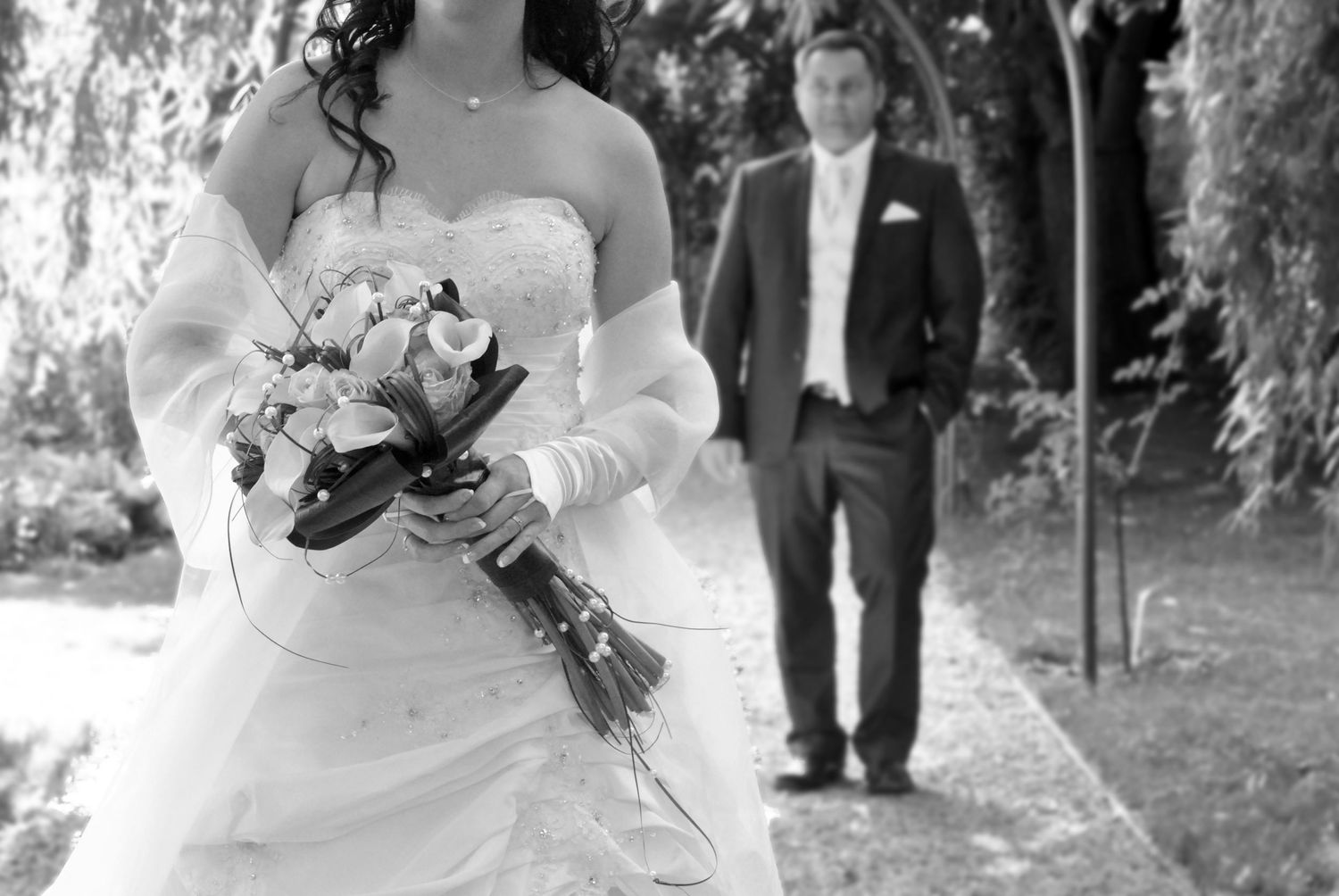 Black and white photo of bride holding bouquet with groom in background.