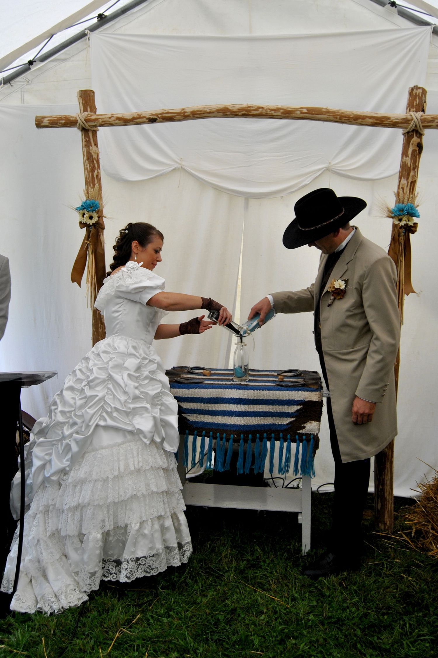 Bride and groom in western attire perform a sand unity ceremony under a rustic wooden arch at an outdoor wedding.