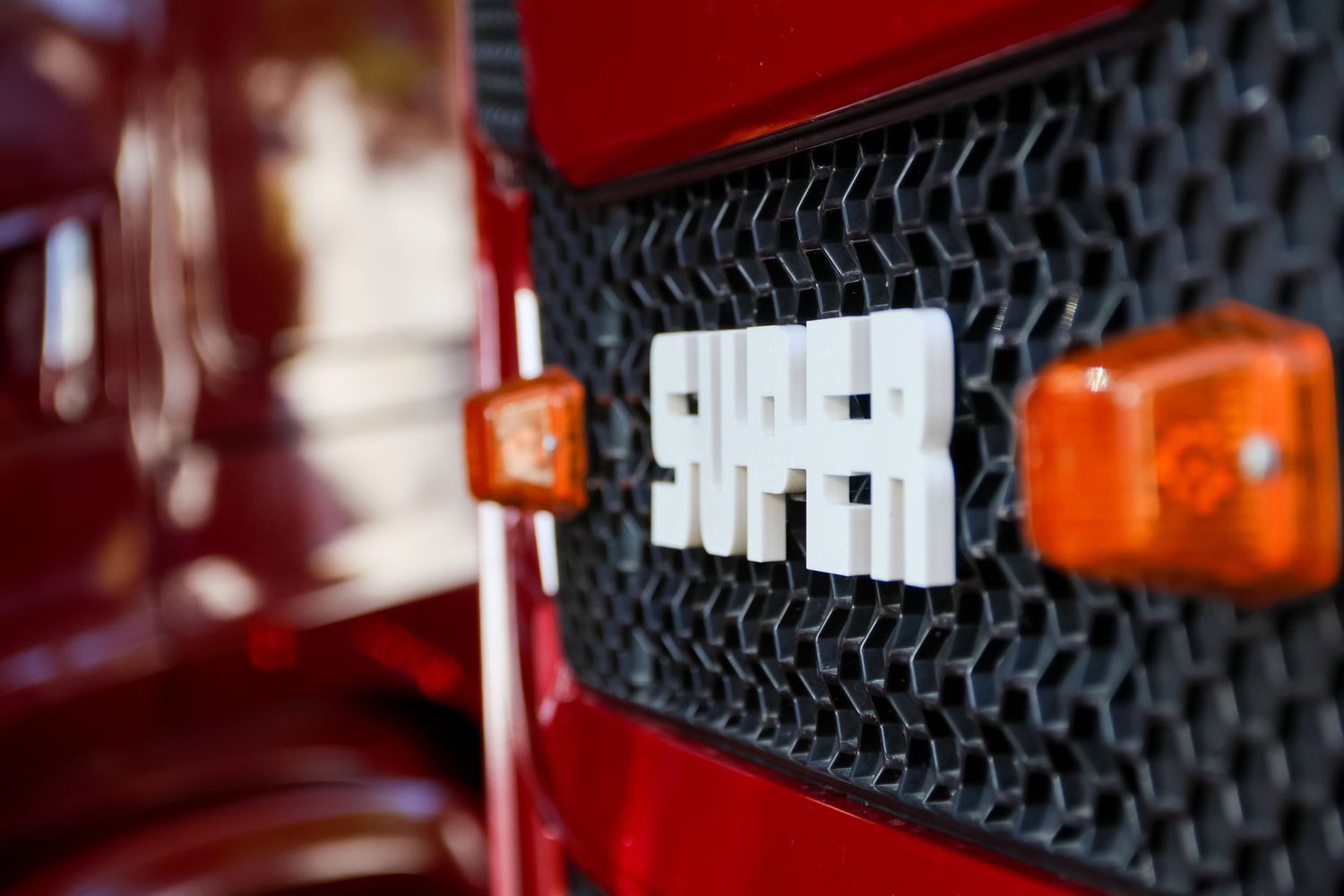 Close-up of a red truck grille featuring a chrome SUPER badge flanked by amber turn signal lights.