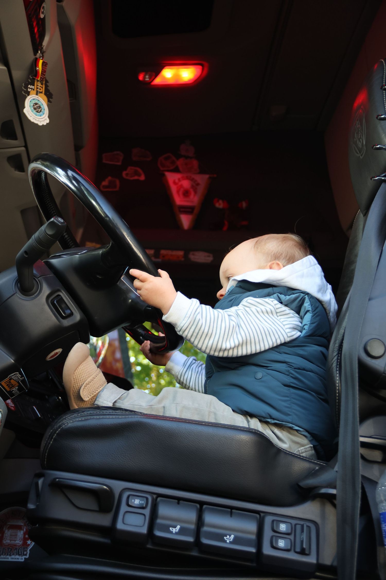 Baby sitting in a truck drivers seat reaching for the steering wheel.