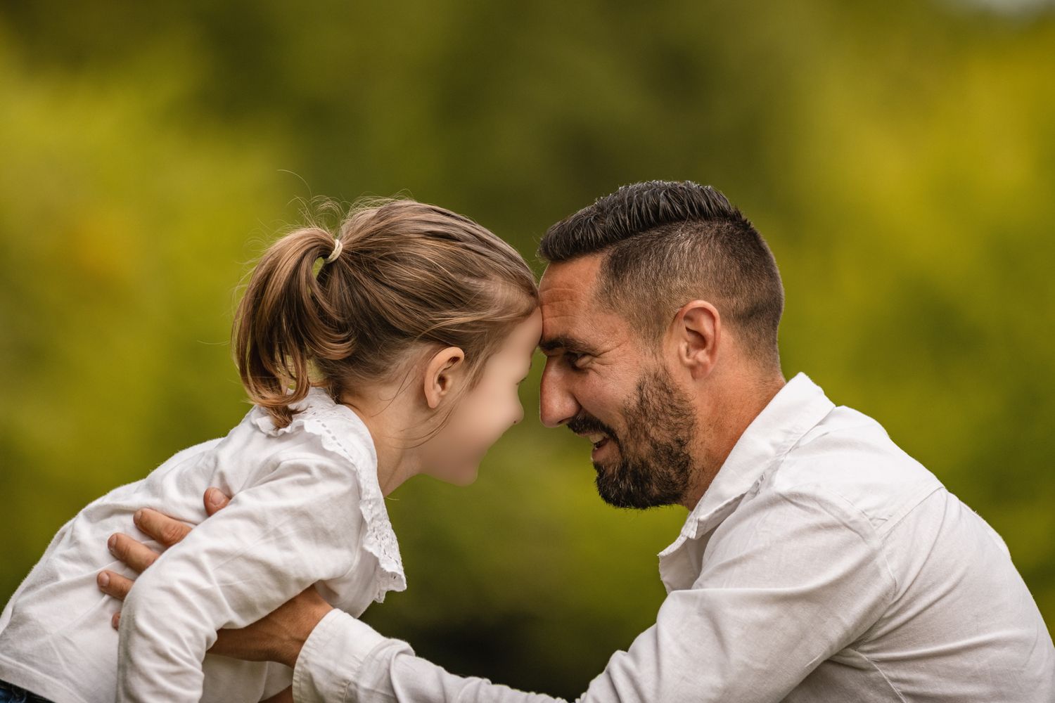 A father and young daughter touch foreheads affectionately outdoors, sharing a tender moment with green foliage behind them.