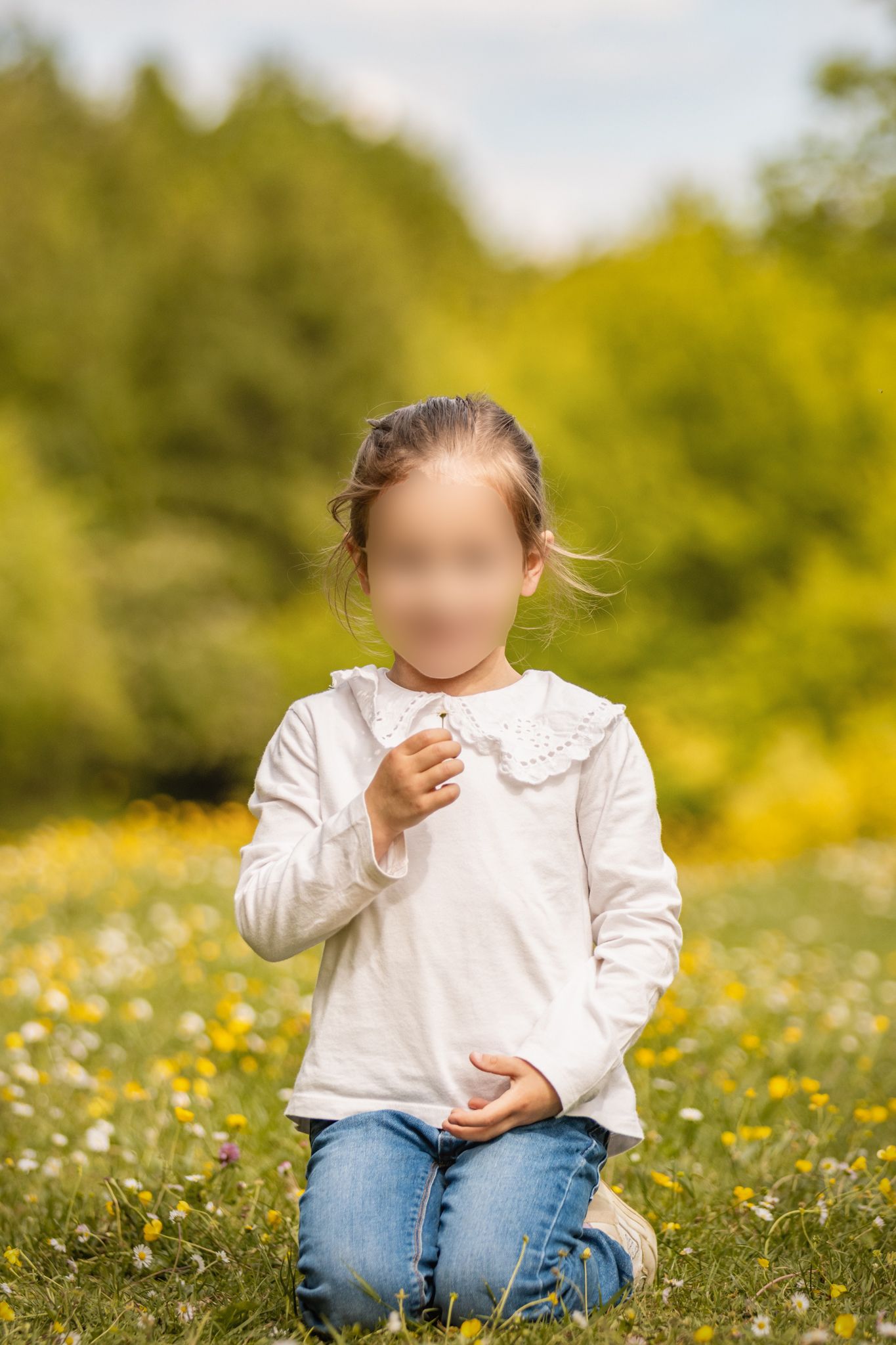 Young girl kneeling in a sunny meadow filled with yellow and white wildflowers, wearing a white top and jeans.
