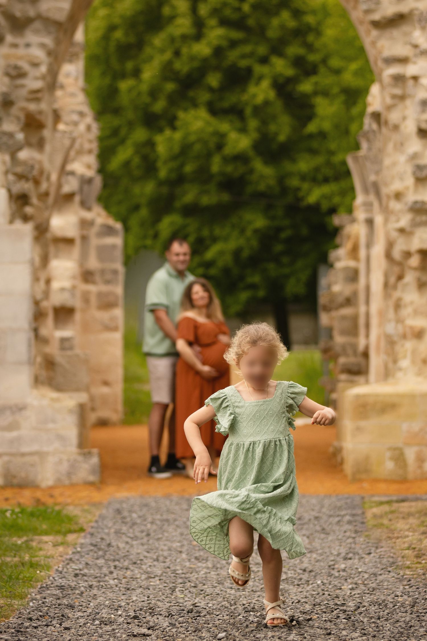 Young girl in green dress runs toward camera as couple stands under stone arch in lush garden setting.