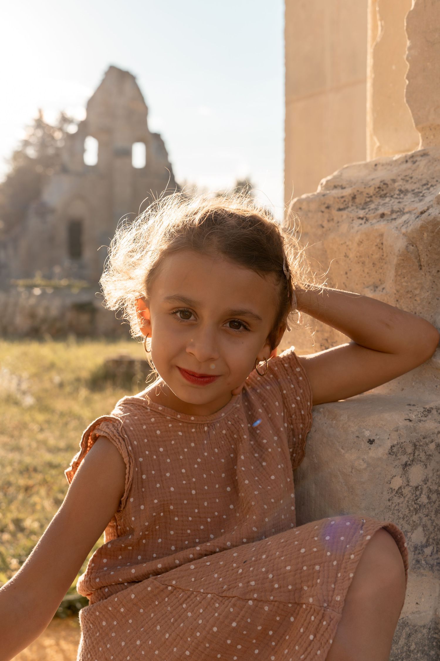 Young girl in polka dot dress leaning against ancient stone ruins at golden hour.