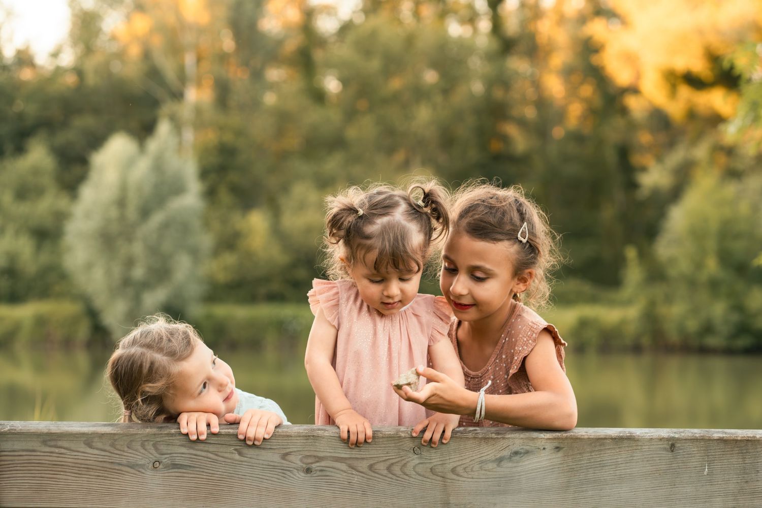 Three young sisters lean over a wooden dock railing near a serene lake in autumn.