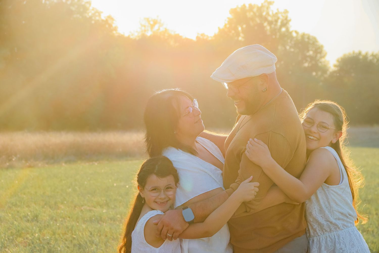 A joyful family of four shares a warm embrace in a sunlit golden field at sunset.