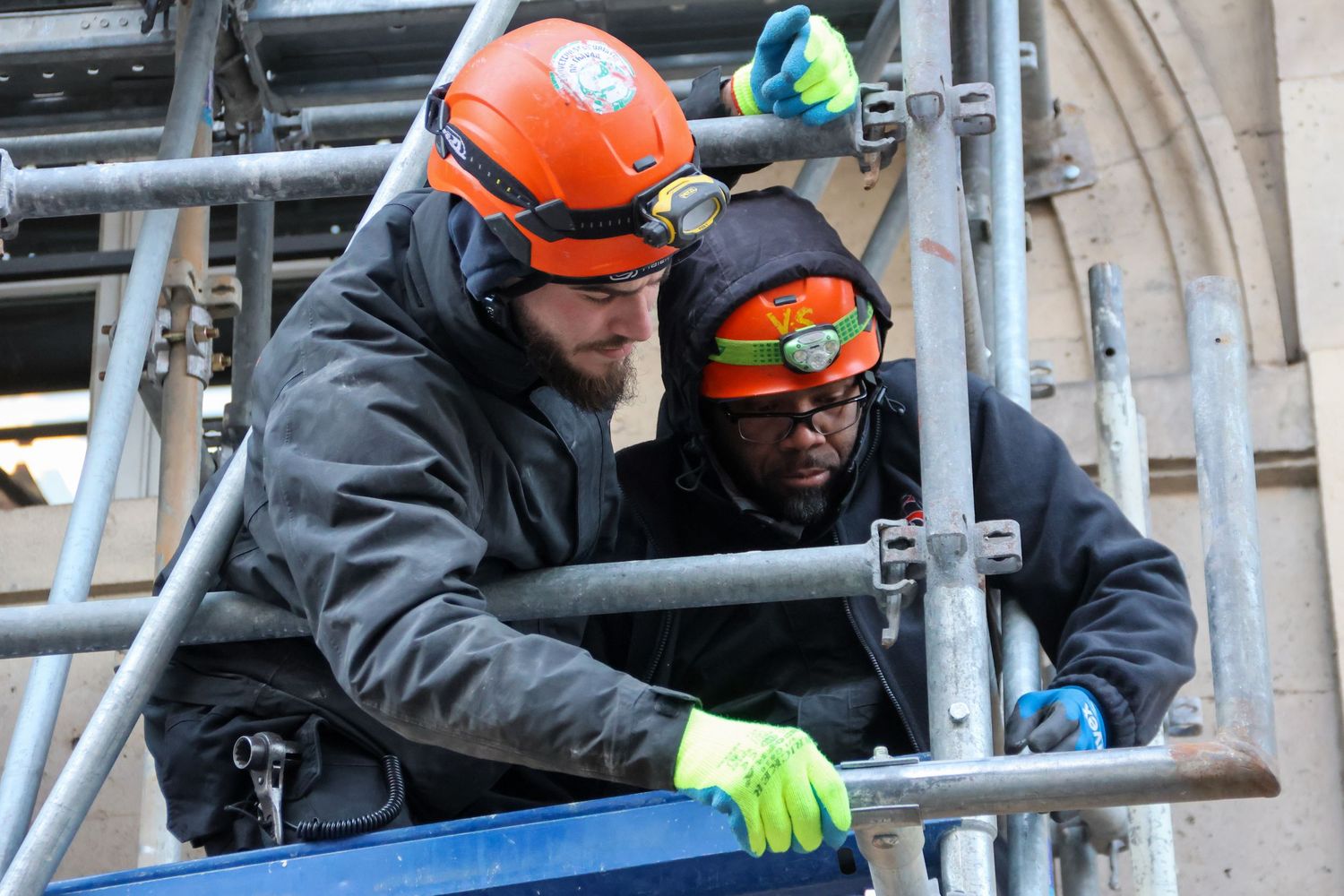 Two construction workers in helmets assembling scaffolding on a building exterior.
