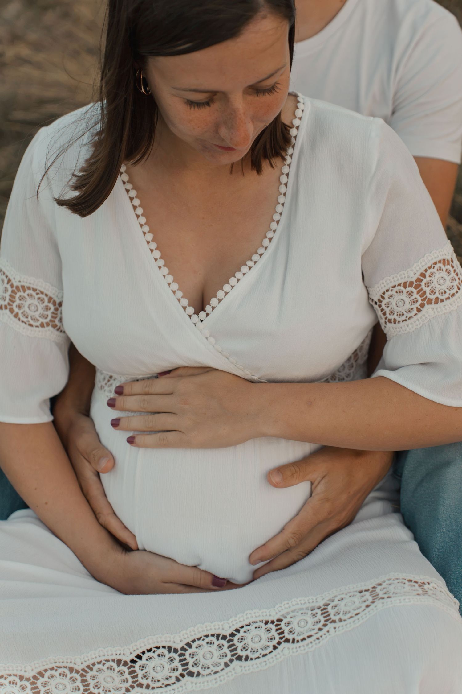 Pregnant woman in white lace dress cradling baby bump with partner outdoors during maternity photo session.