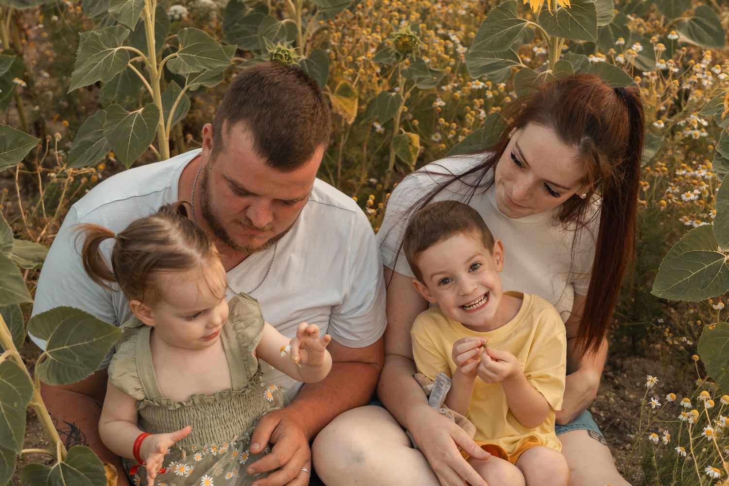 A happy young family with two toddlers sitting together outdoors among sunflower plants in warm golden light.