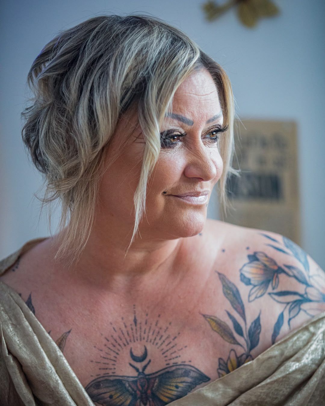 Woman with blonde updo, blue eye makeup, and floral chest tattoos smiles over her shoulder, wearing a gold top.